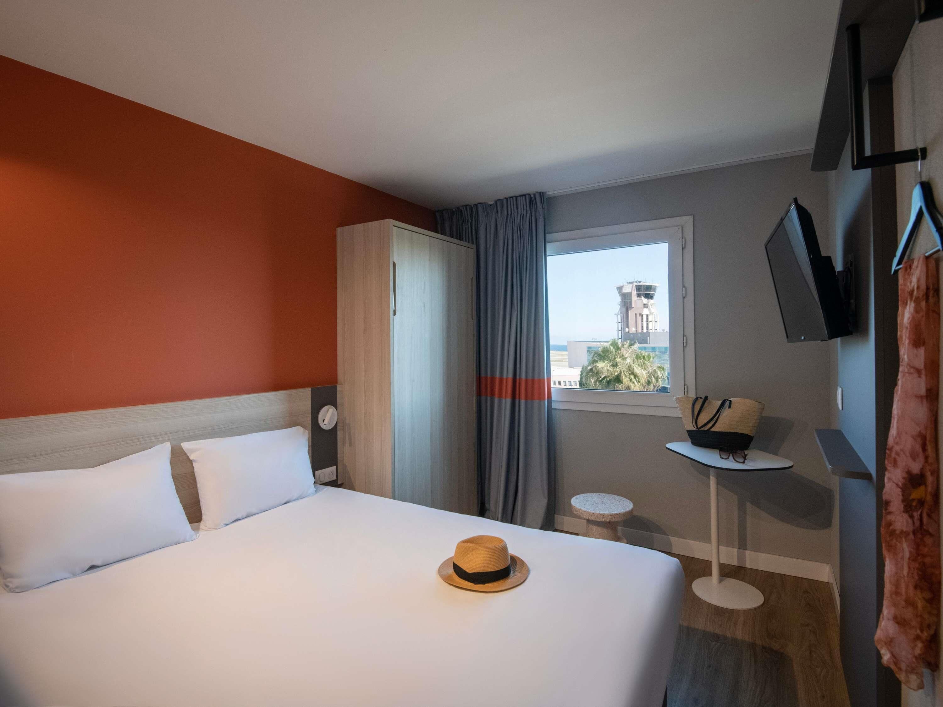A hotel room with a bed, a small table, a TV, and a window with a view of a building and palm trees.