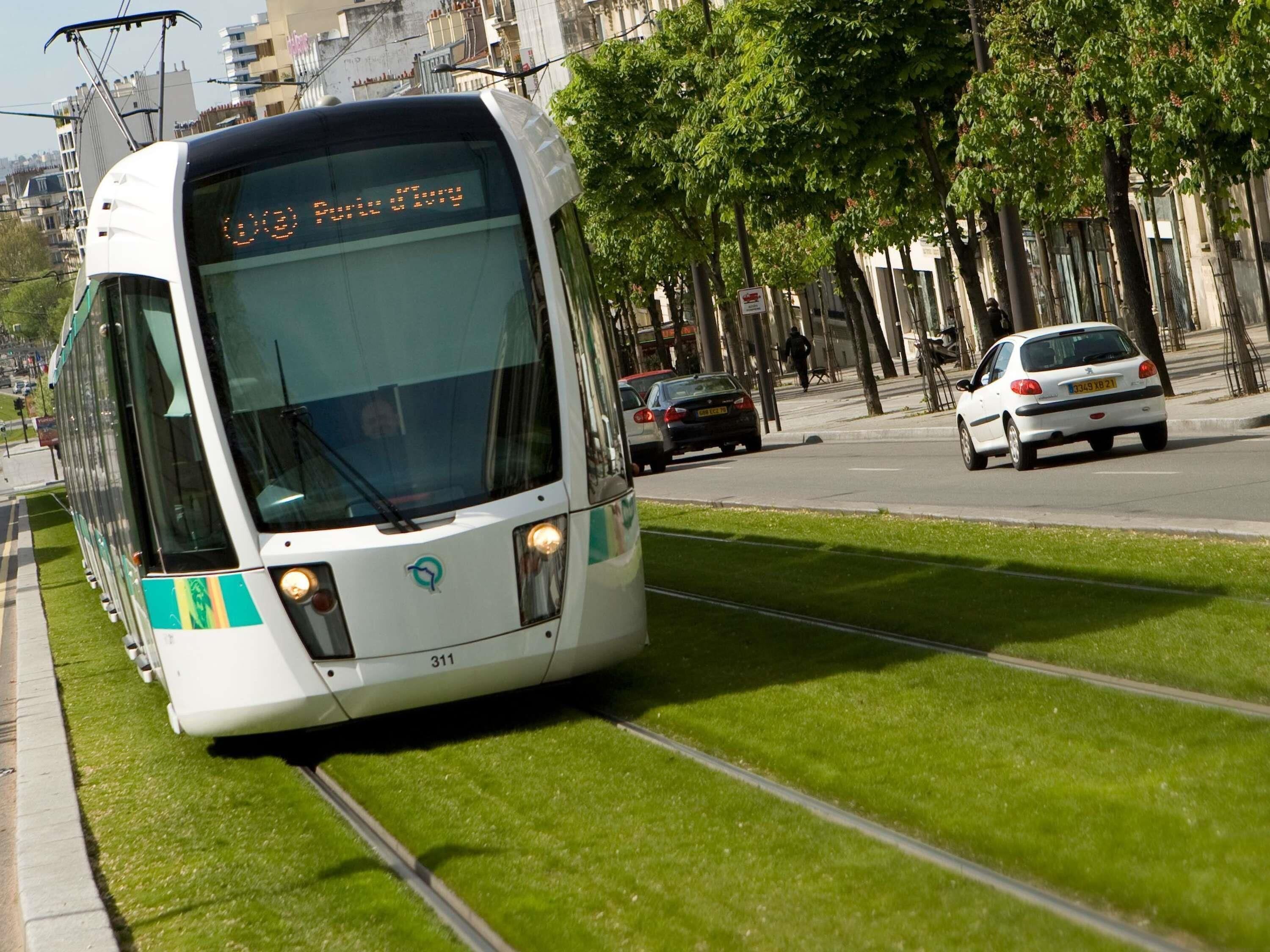 Een tram rijdt over rails, met een digitaal display, omgeven door stadsgebouwen en bomen.