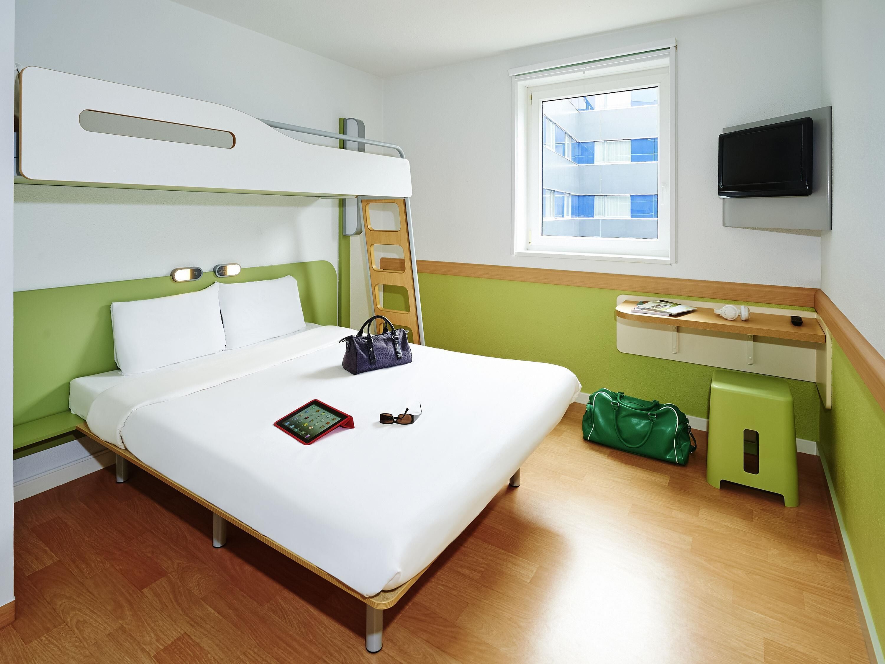 Una habitación de hotel con literas, una mesita de noche con televisor, una ventana con vistas a edificios y un bolso verde en el suelo.