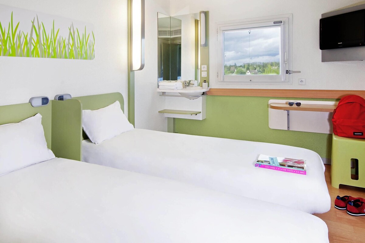 Une chambre d’hôtel avec deux lits, un miroir, une fenêtre, une télévision et une petite table sur laquelle se trouvent un magazine et une paire de chaussures.