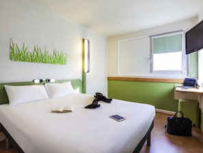 Habitació d'hotel amb un llit, una taula de treball amb un ordinador portàtil, una finestra i una obra d'art a la paret.