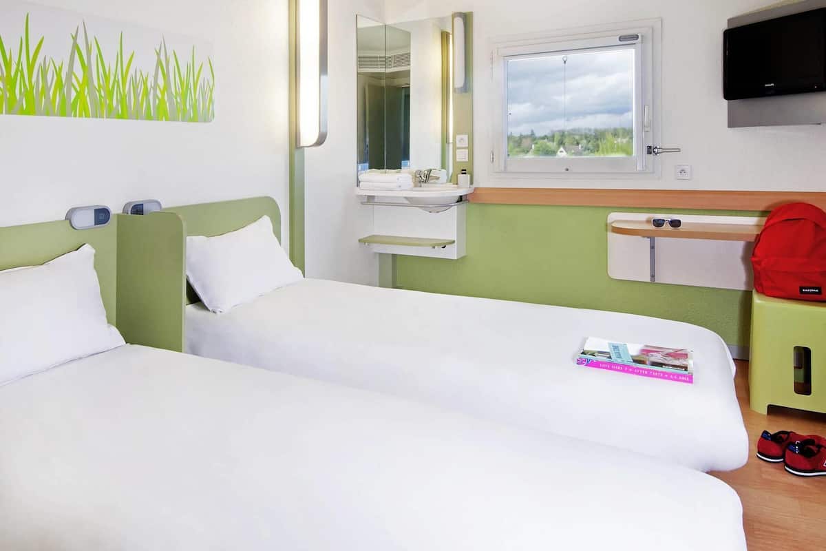 Een hotelkamer met twee bedden, een spiegel, een raam, een televisie en een klein tafeltje met een tijdschrift en een paar schoenen.