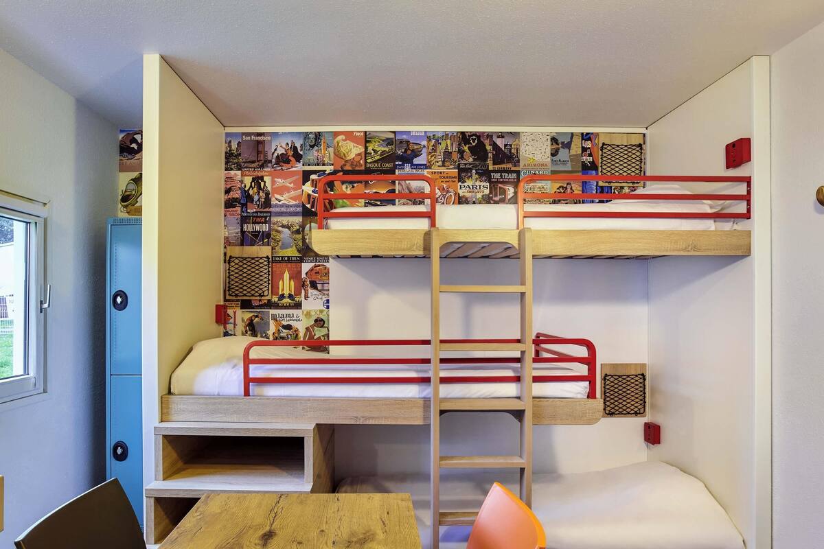 A bunk bed room with a desk and chair, a window, and a wall decorated with travel posters.