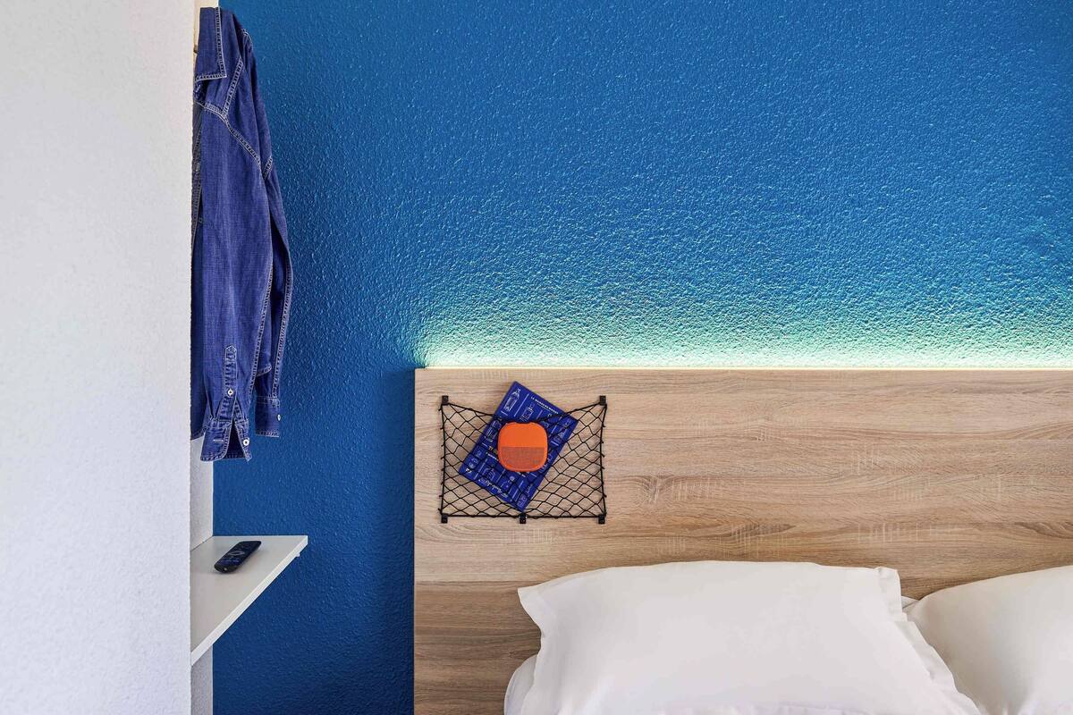 A bedroom with a wooden headboard, a blue textured wall, a hanging denim jacket, a small shelf with a remote control, and a neatly made bed with white pillows.
