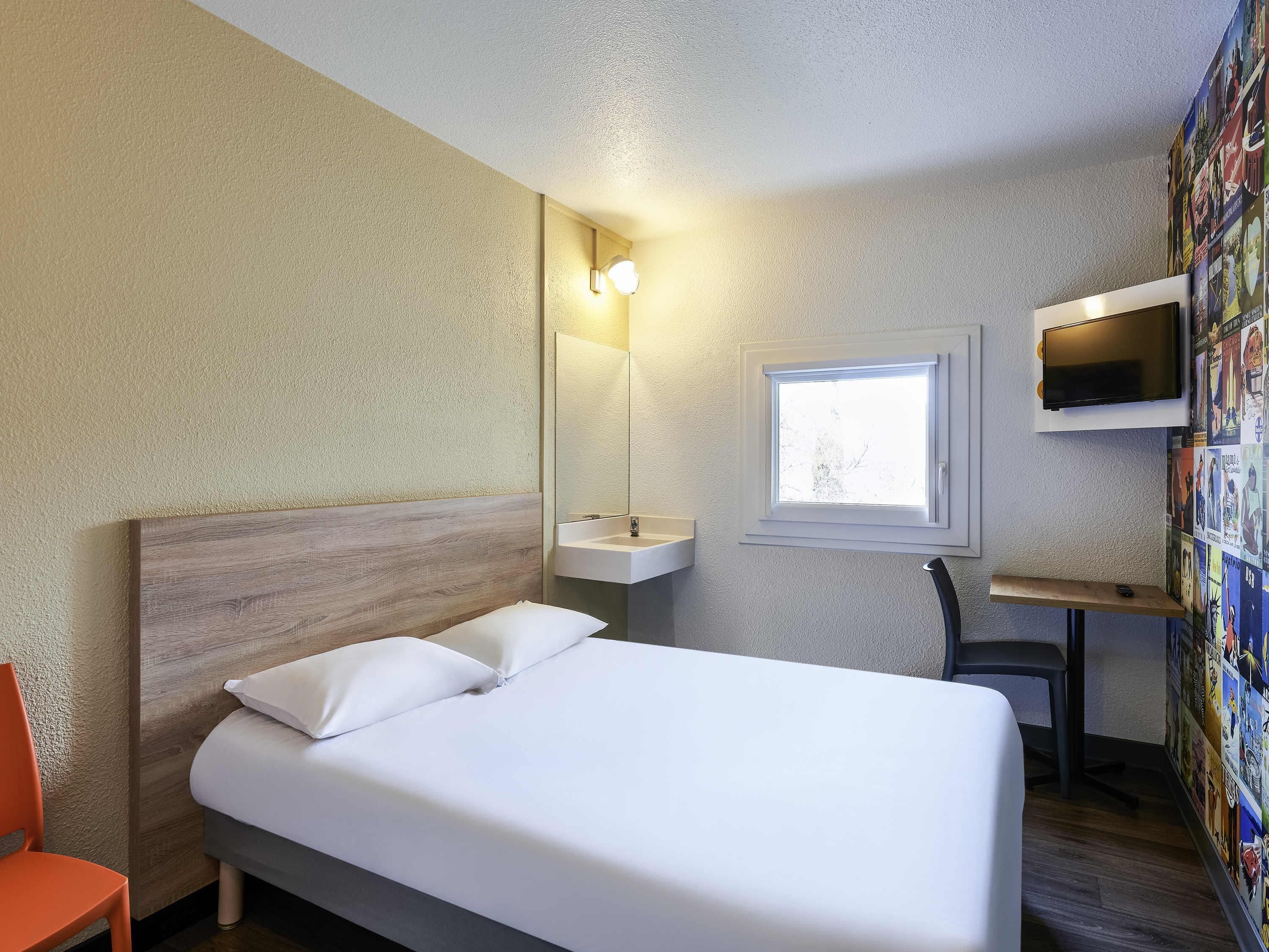 Ein Hotelzimmer mit einem Bett, einem Schreibtisch, einem Stuhl, einem an der Wand befestigten Fernseher, einem Fenster und einer Wand mit einem Poster.