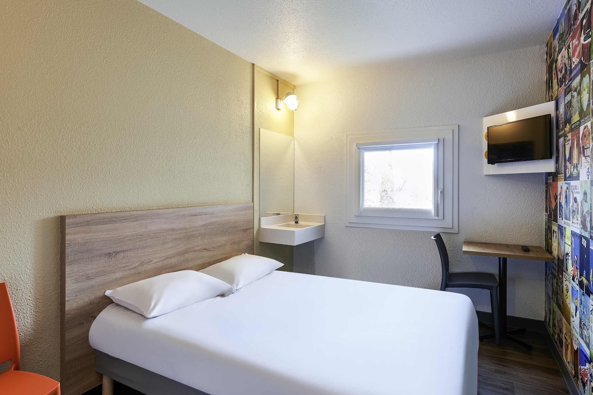 A hotel room with a bed, a desk, a chair, a wall-mounted TV, a window, and a wall with a poster.