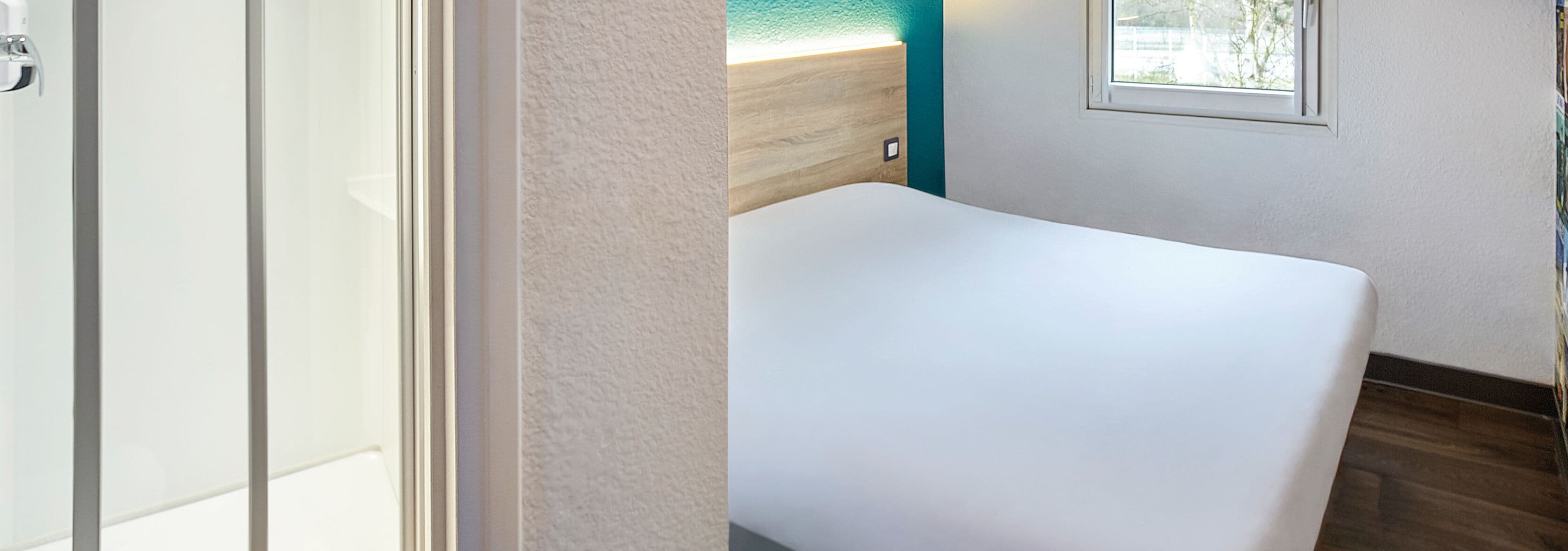 A hotel room with a white bed, a wooden headboard, a small window with a view, and a wall-mounted light fixture.
