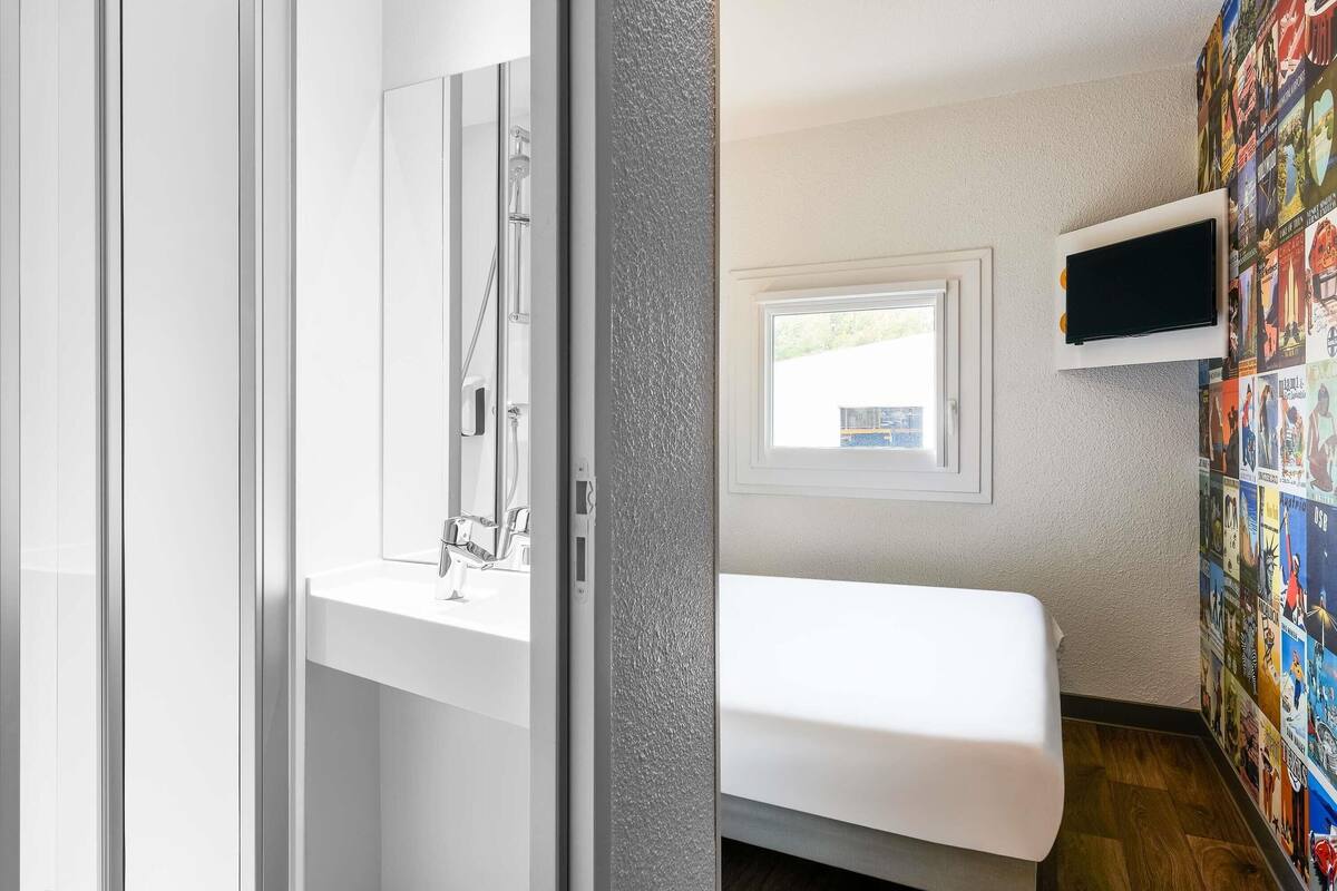 A room with a bed, a TV mounted on the wall, a window, and a bathroom with a shower.