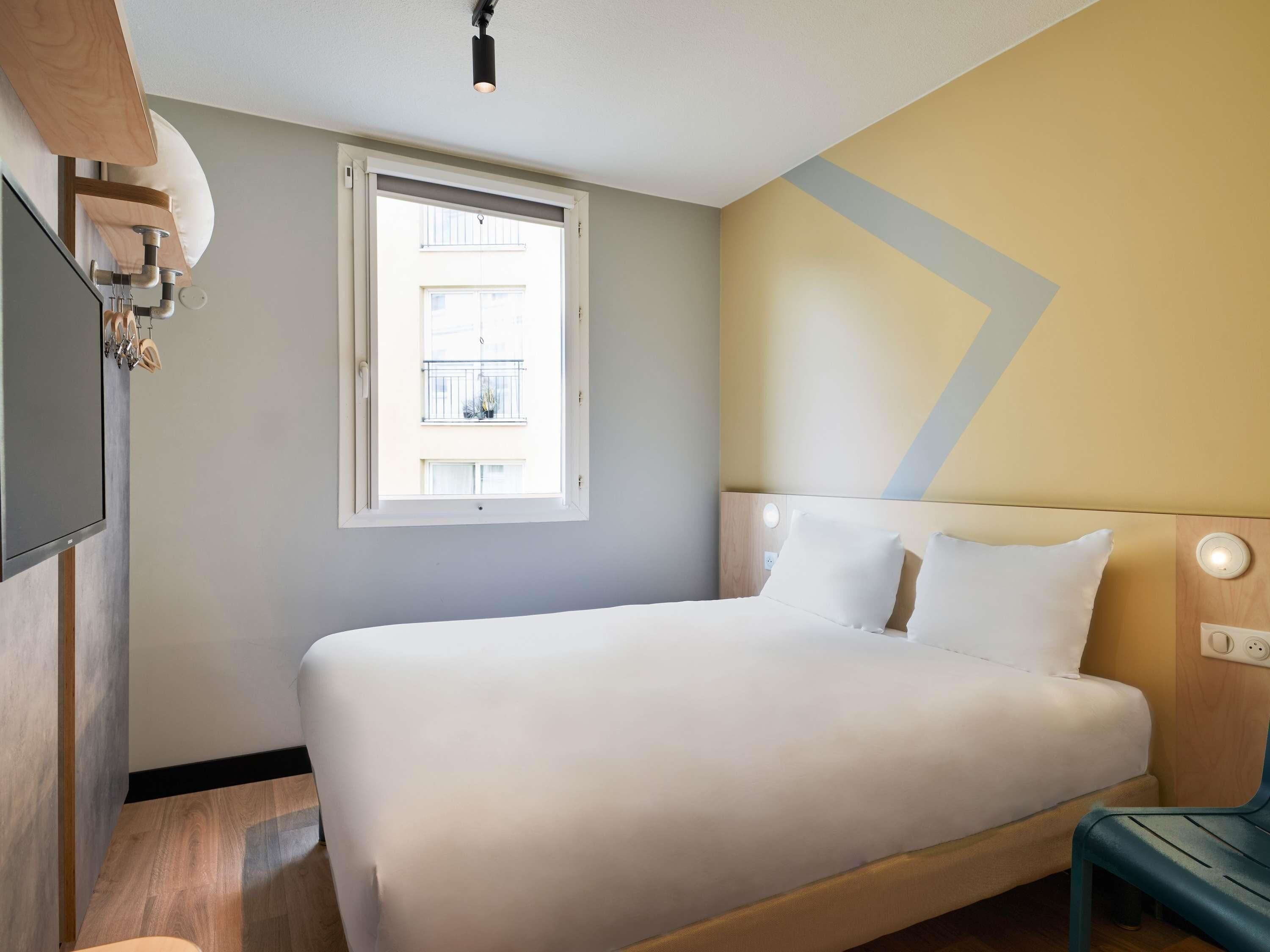 Een hotelkamer met een bed, een raam, een wandlamp en een houten hoofdeinde.