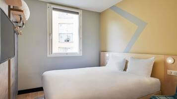 A hotel room with a bed, a window, a wall-mounted light, and a wooden headboard.