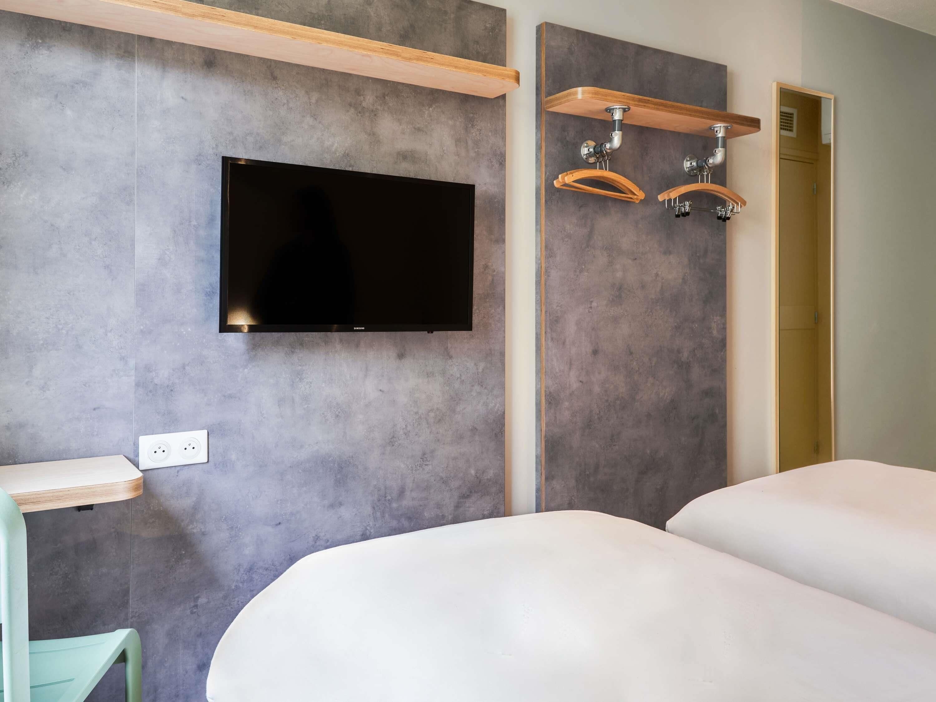 Een hotelkamer met een wandgemonteerde flatscreen-tv, een houten plank met haken, een tafeltje en een stoel.