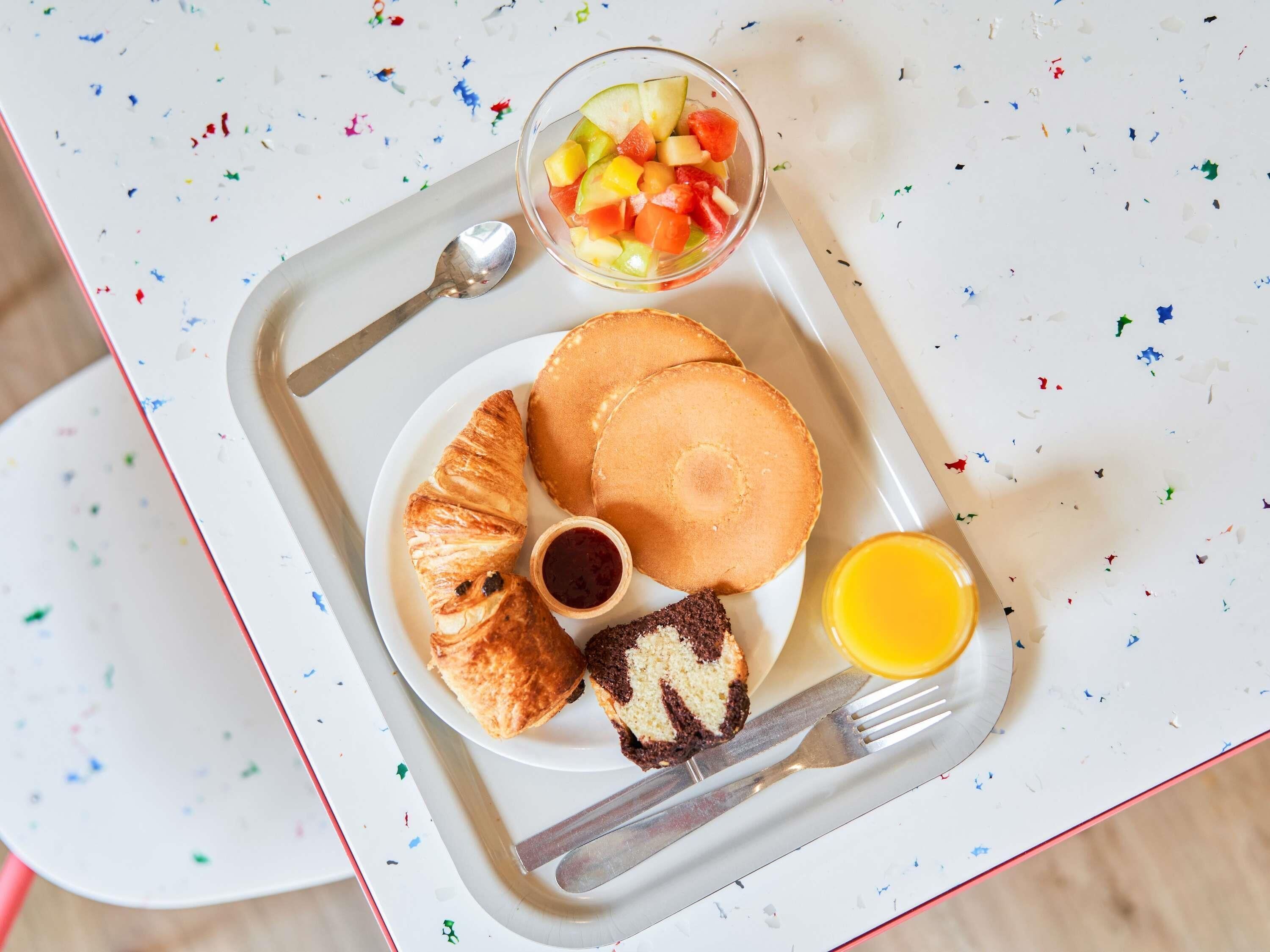 Un plat de petit esmorzar amb crepes, un croissant, un muffin, una amanida de fruita i un got de suc d'aranja.