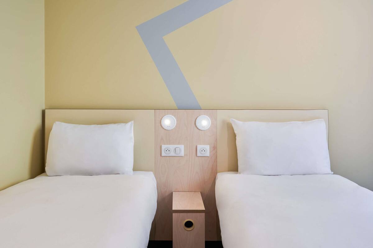 A hotel room with two beds, a wooden headboard, two wall-mounted light switches, and a small wooden nightstand.