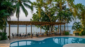 A poolside area with a covered seating area and tables, surrounded by palm trees and a view of the sea. - Banana Palms Hotel (Livingston)