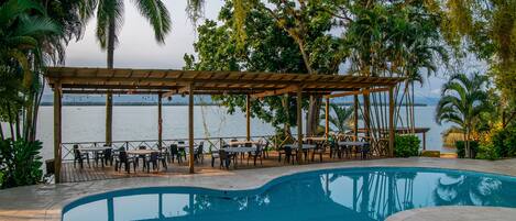 Zona junto a la piscina con un área de estar techada y mesas, rodeada de palmeras y vista al mar.