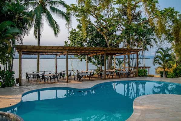 A poolside area with a covered seating area and tables, surrounded by palm trees and a view of the sea.