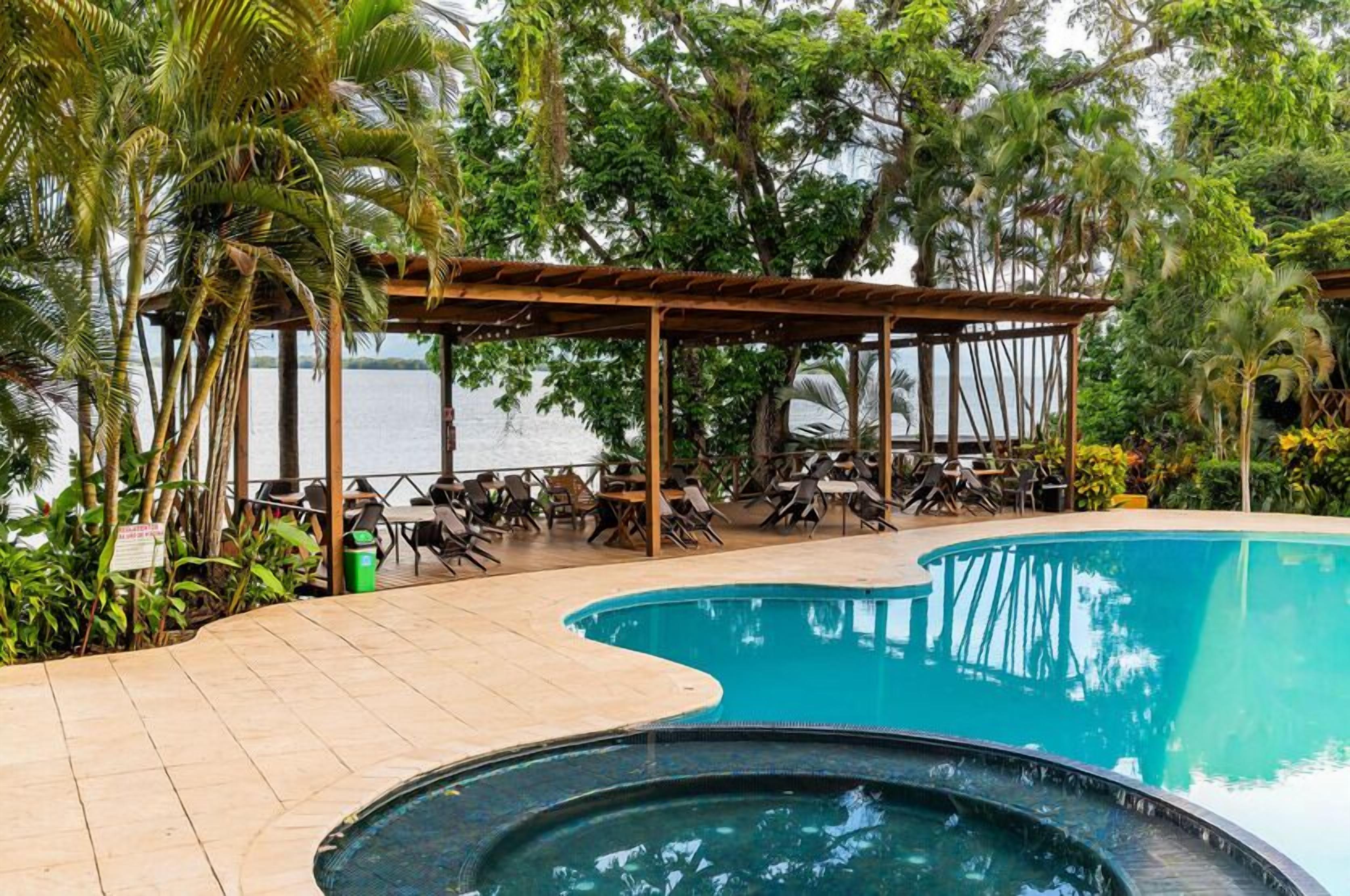 Una piscina con borde curvo, rodeada de sillones de descanso y una zona cubierta para sentarse, con vista al agua.