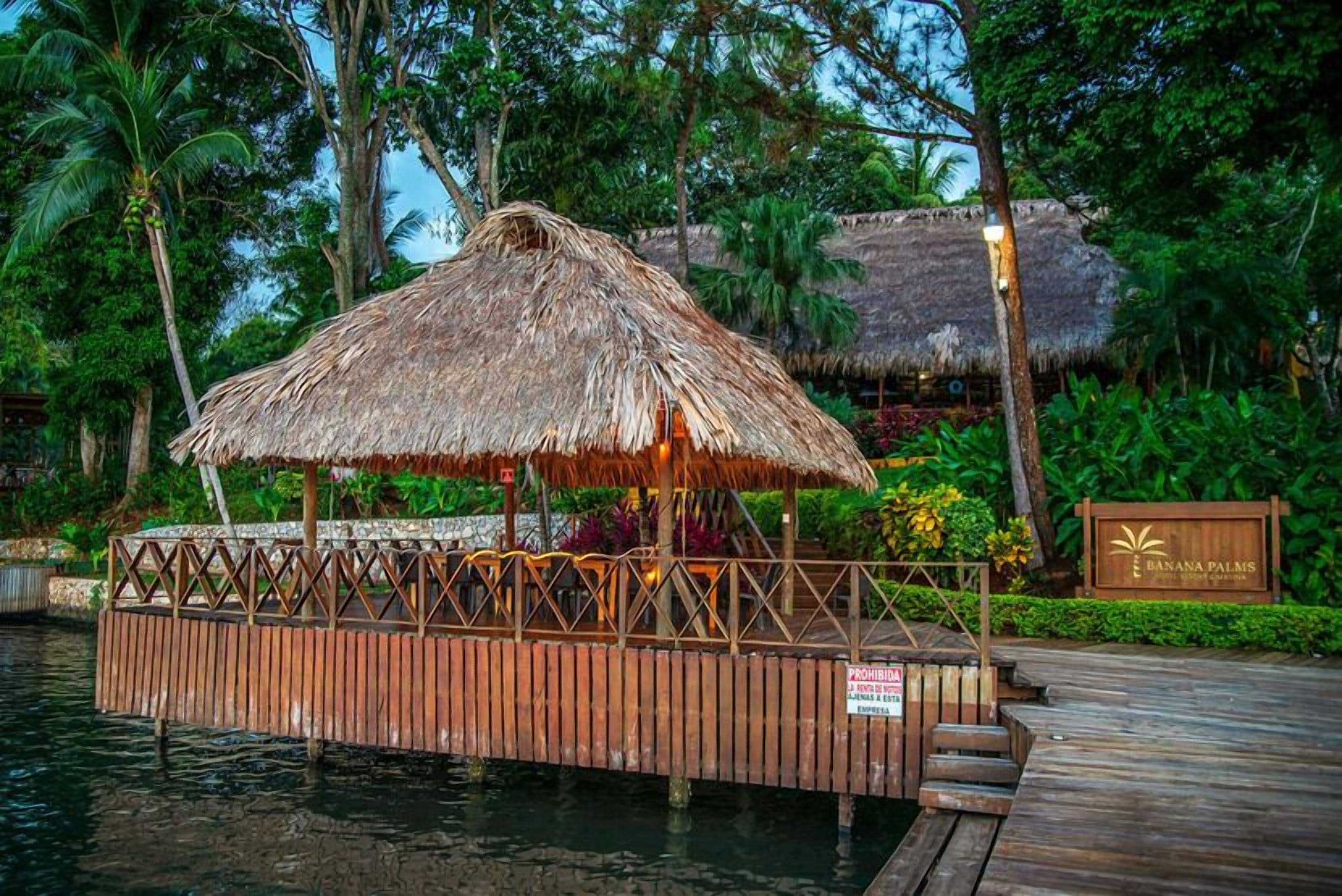 A wooden dock with a thatched-roof structure, surrounded by lush greenery and a sign indicating a resort named 'Banana Palms.'