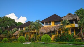 A row of yellow buildings with thatched roofs, surrounded by lush greenery and palm trees. - Banana Palms Hotel (Livingston)
