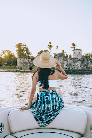 A person sitting on a boat with a view of a historical stone structure and palm trees in the background.