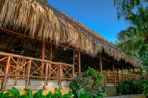 Una estructura de techo de paja con vigas de madera y un muro de celosía, rodeada de exuberante vegetación tropical.