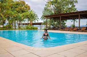 Una piscina con una persona en el agua, rodeada de palmeras y una zona cubierta con sillas de descanso.