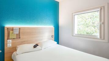 A hotel room with a bed, a headboard with a charging station, a window with a view of greenery, and a wall with a blue accent.