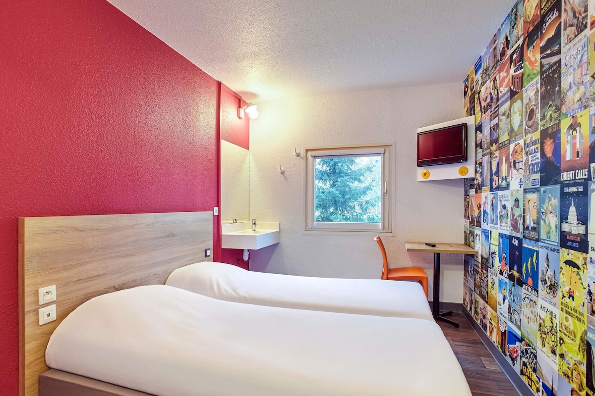 Une chambre d’hôtel équipée d’un lit, d’un bureau, d’une télévision et dont le mur est décoré d’affiches de voyage.