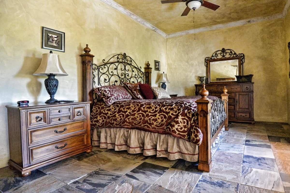 A bedroom with a four-poster bed, a dresser with drawers, a nightstand, a ceiling fan, a mirror, and a window with curtains.
