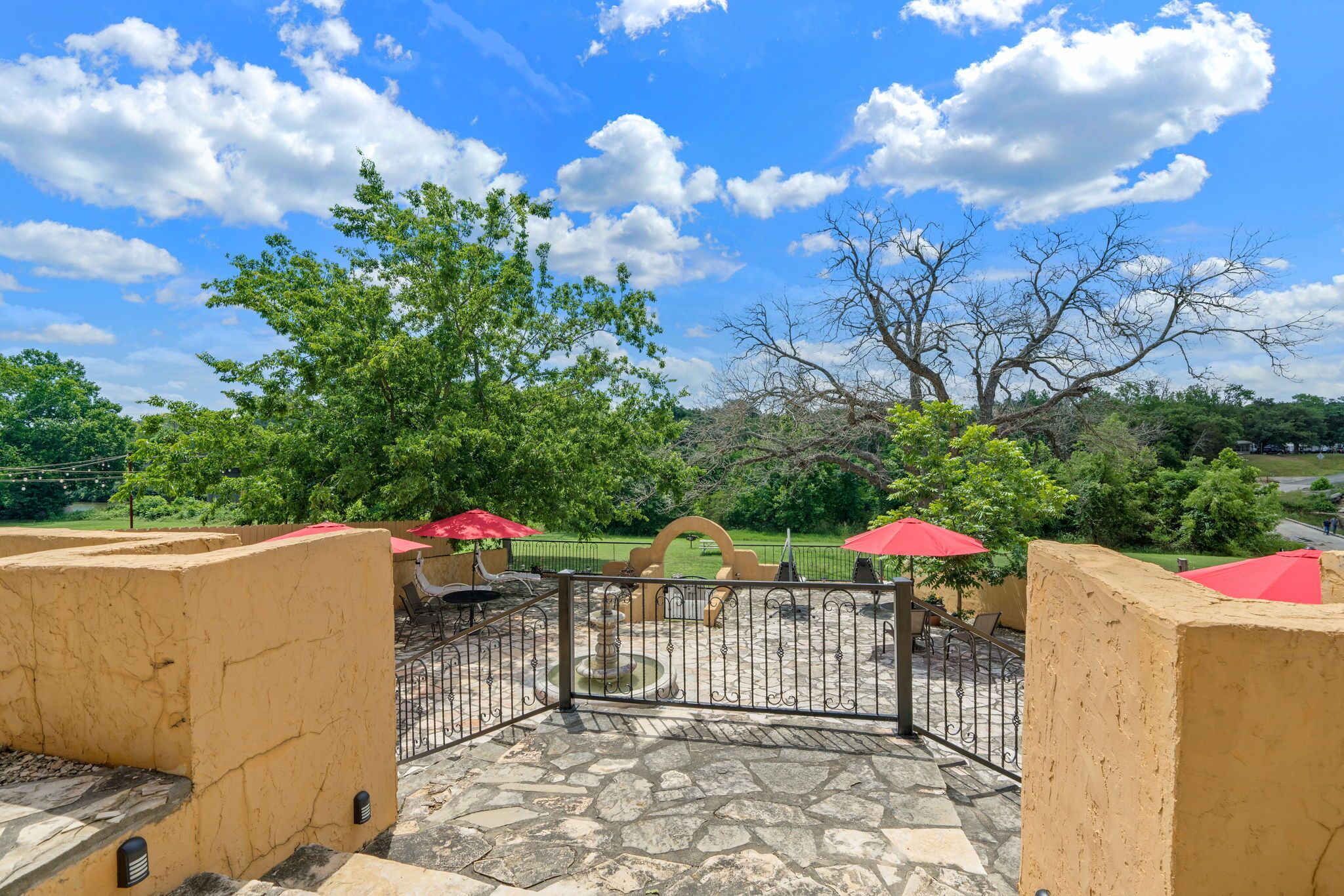 An outdoor patio area with stone flooring, red umbrellas, and a view of a park.