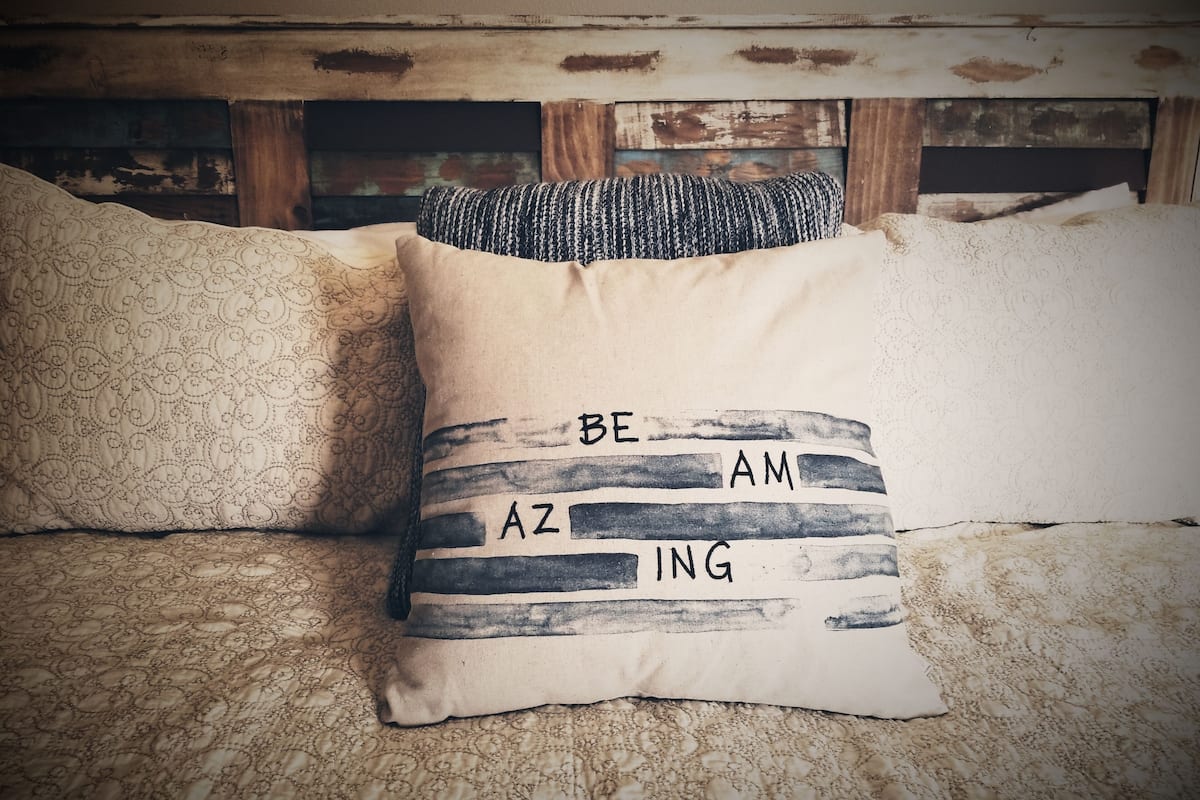 A bed with patterned bedding and a pillow with the text "BE AM AZ ING".