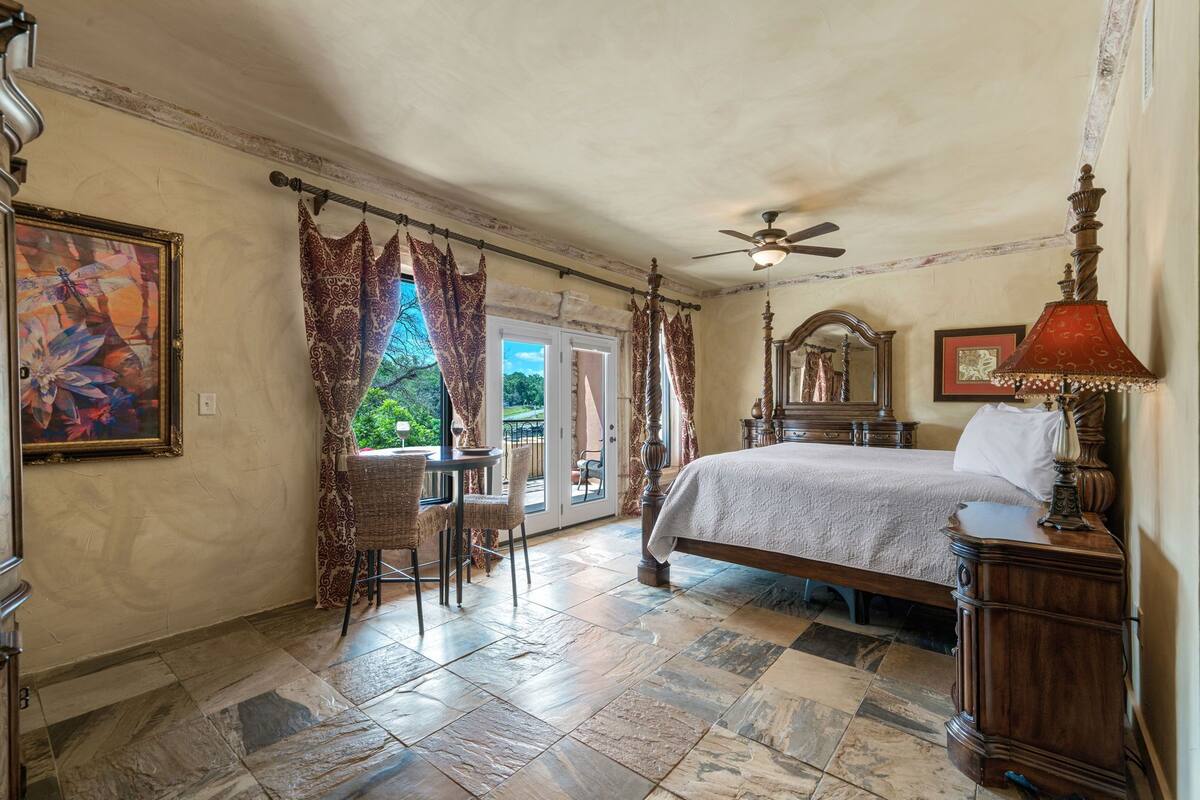 A bedroom with a bed, a ceiling fan, a table with chairs, and a view of the outdoors.