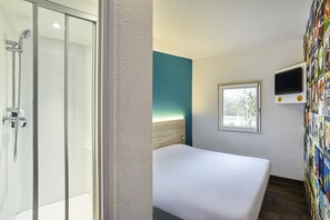 Habitación de hotel con cabina de ducha de vidrio, cama con cabecera de madera, ventana pequeña y televisor montado en la pared.