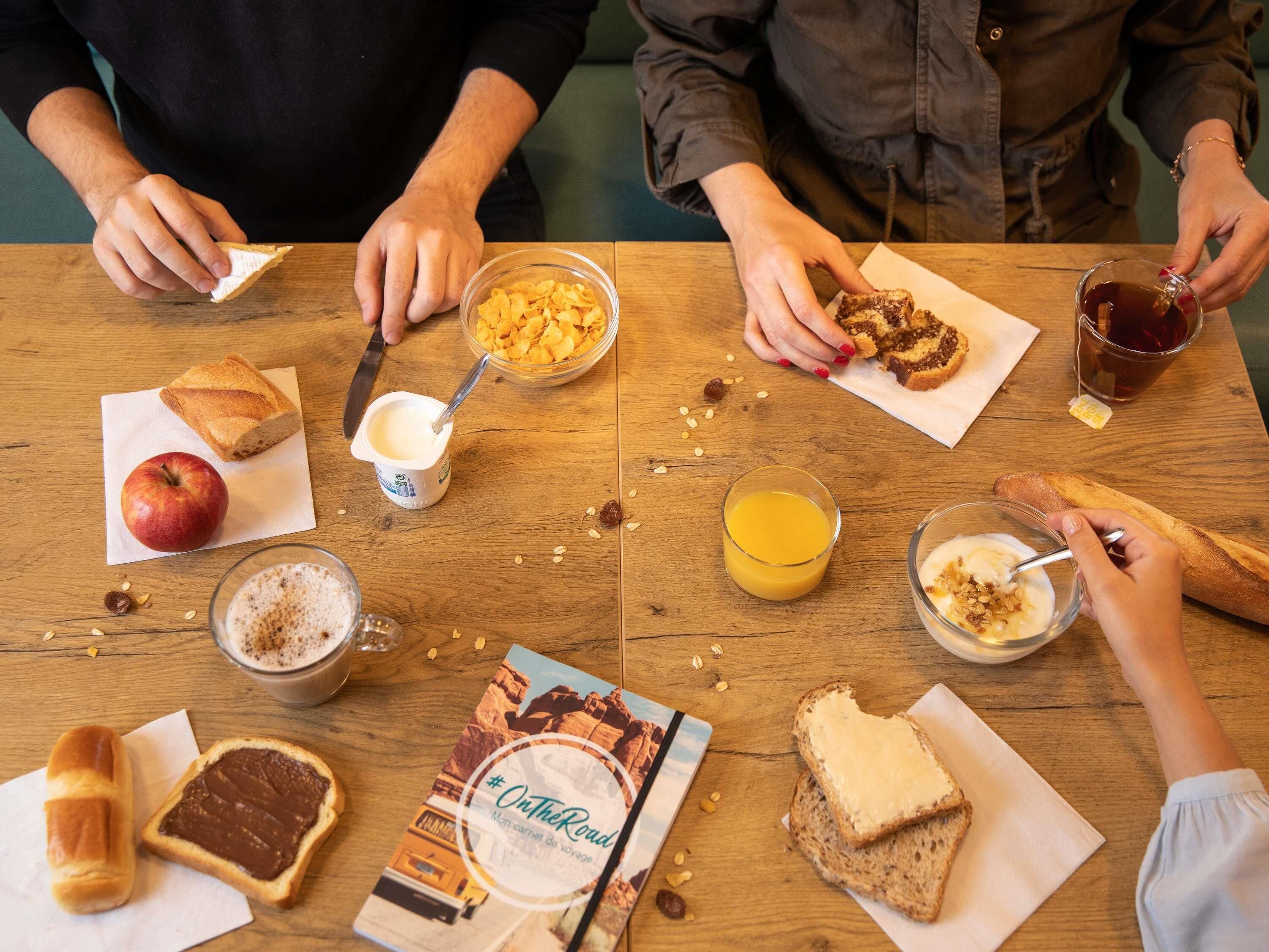 Et træbord med forskellige morgenmadsvarer, herunder brød, müsli, frugt og drikkevarer.
