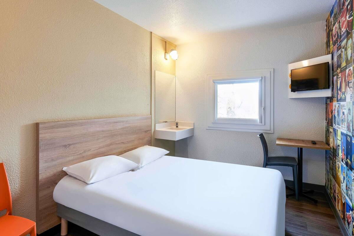 A hotel room with a bed, a desk, a chair, a TV, a window, and a wall with a poster.