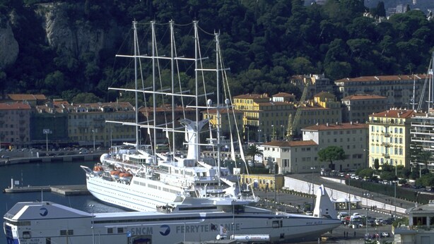 Un paysage urbain côtier avec un grand navire de croisière amarré dans un port, entouré de bâtiments et d’une route où circulent des voitures.