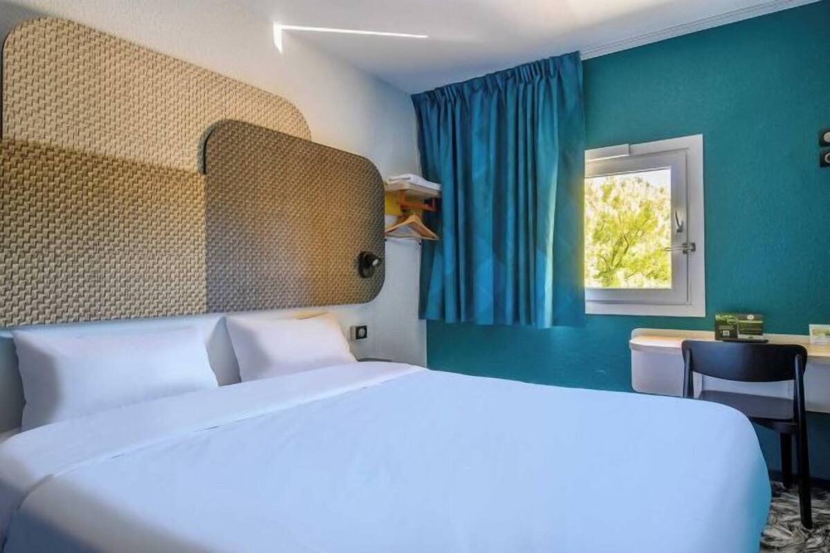 A hotel room with a large bed, a desk, a chair, a window with curtains, and a wall-mounted shelf.