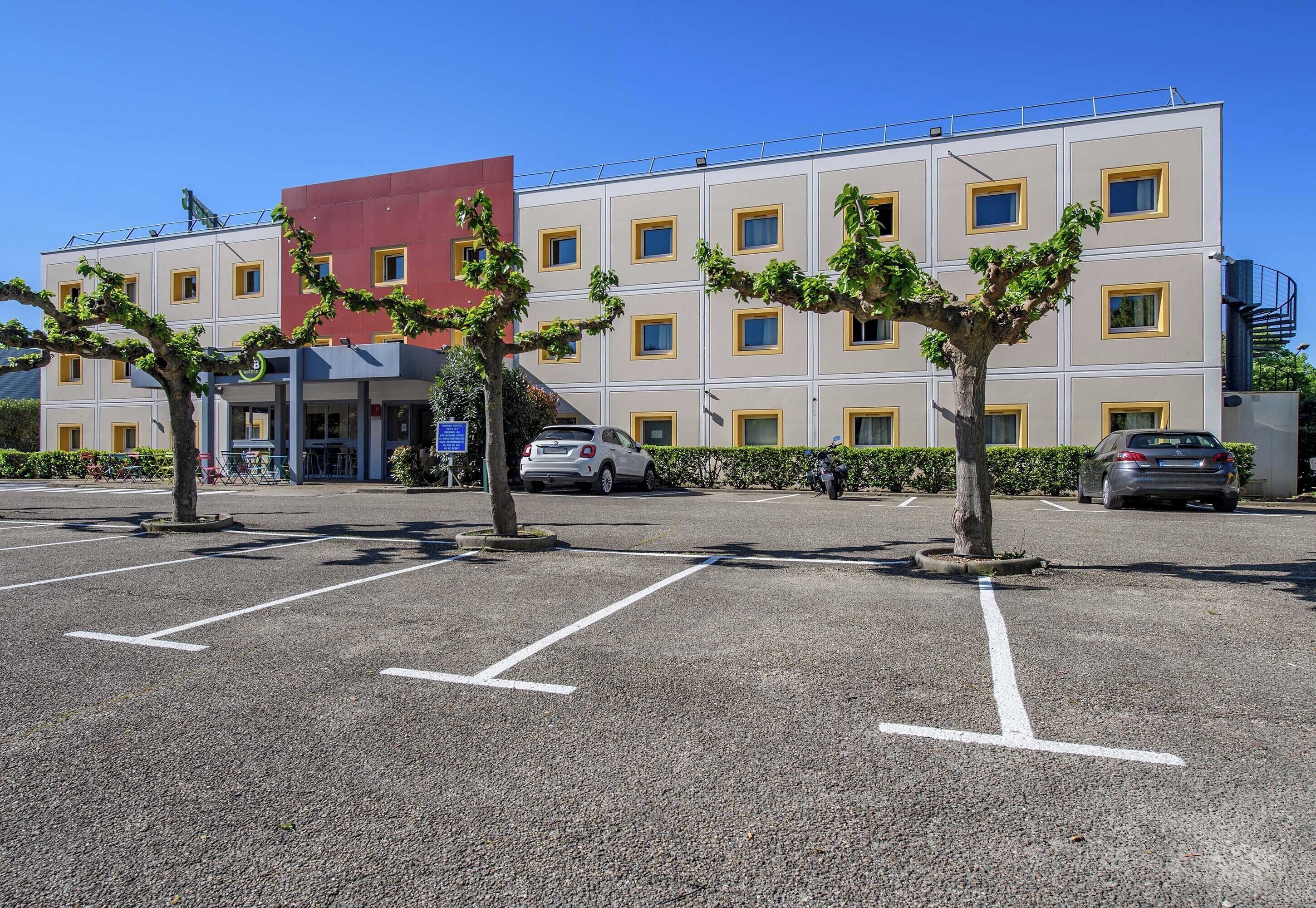 A multi-story hotel with a parking lot, trees, and a red building in the background.