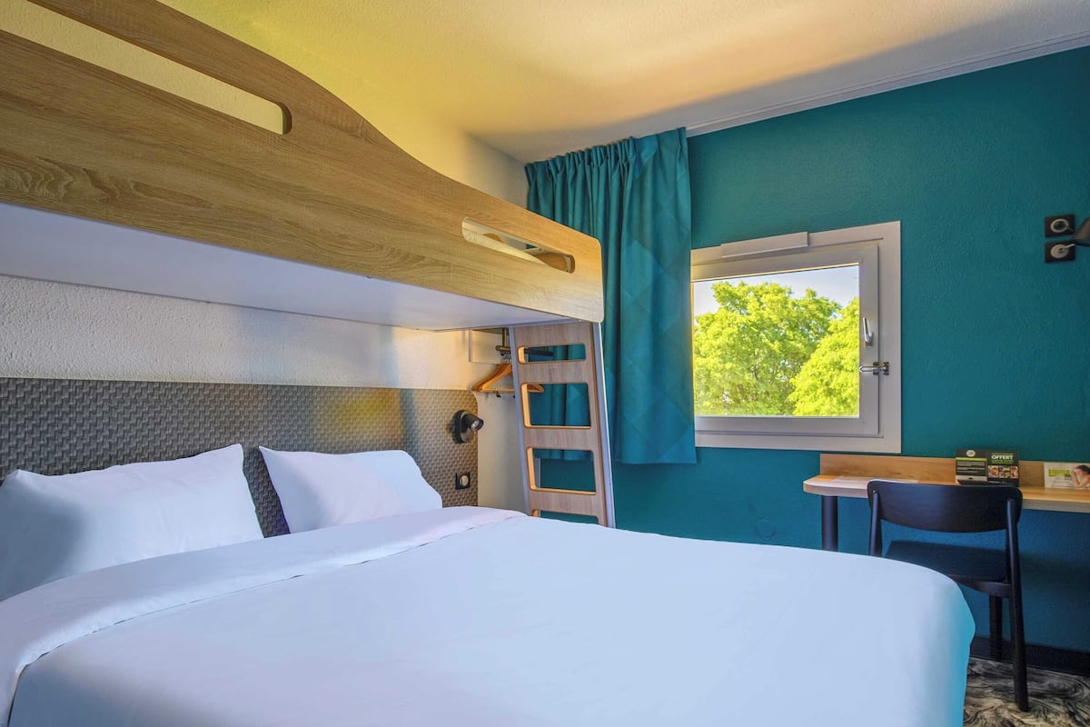 A hotel room with a bunk bed, a desk with a chair, a window with curtains, and a view of trees outside.