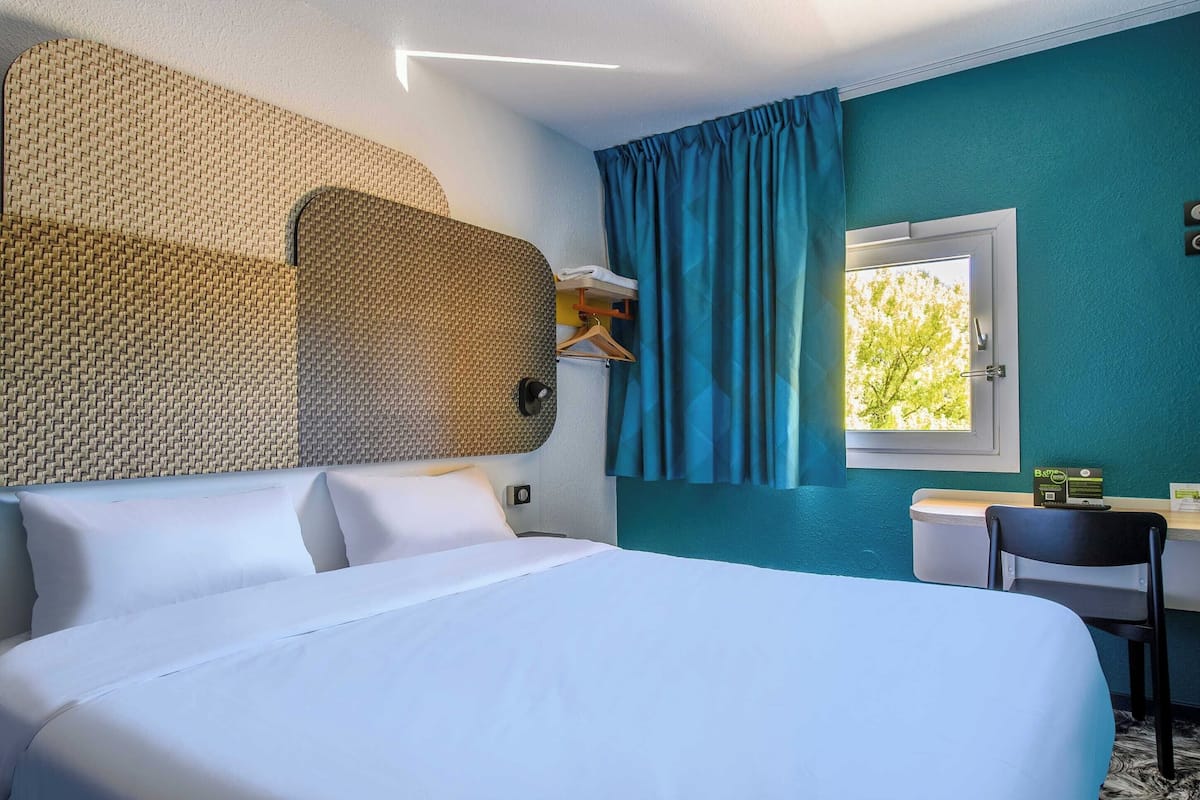 A hotel room with a large bed, a desk with a chair, a wardrobe, and a window with curtains.