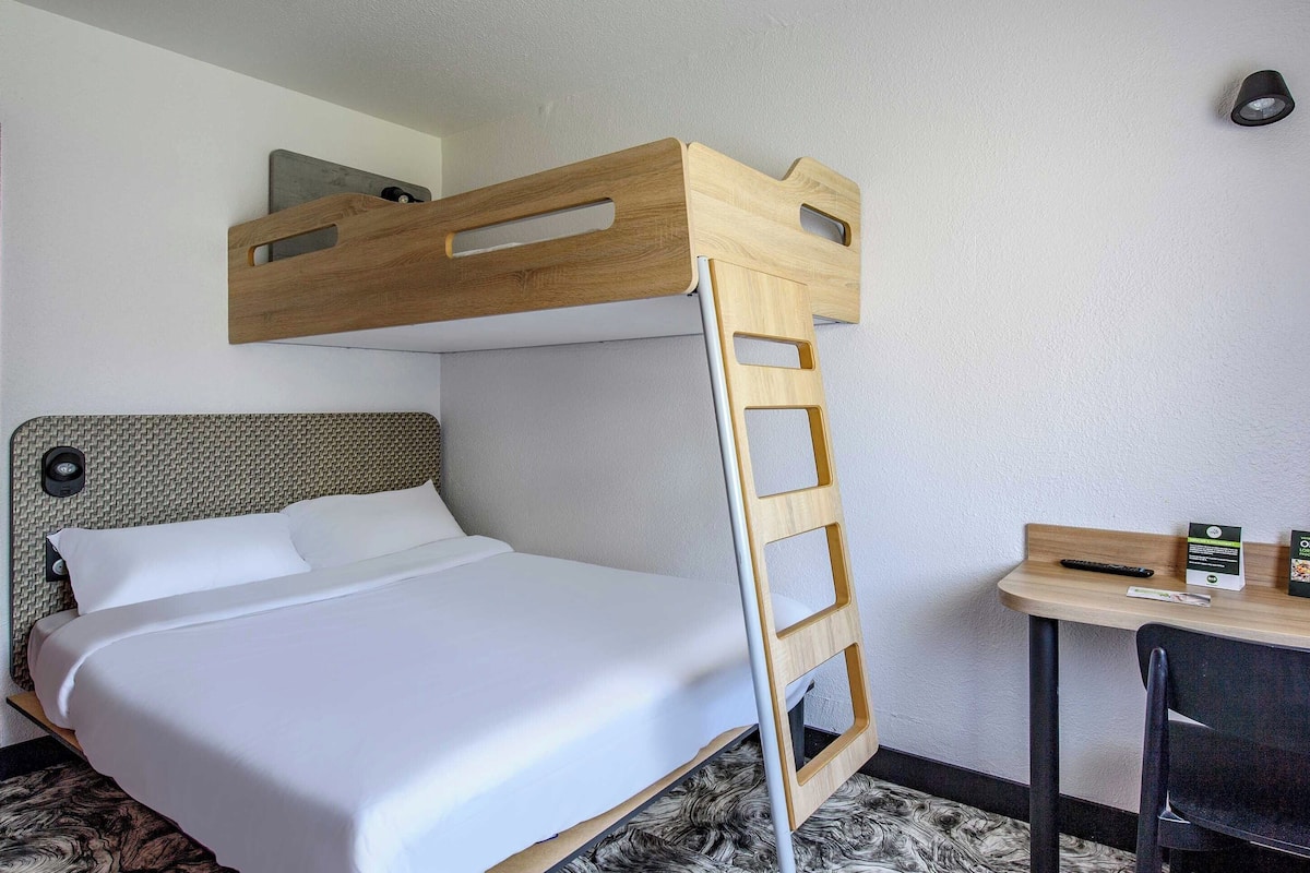 A bunk bed with a wooden ladder, a desk with a chair, and a small table with a remote control and a box.