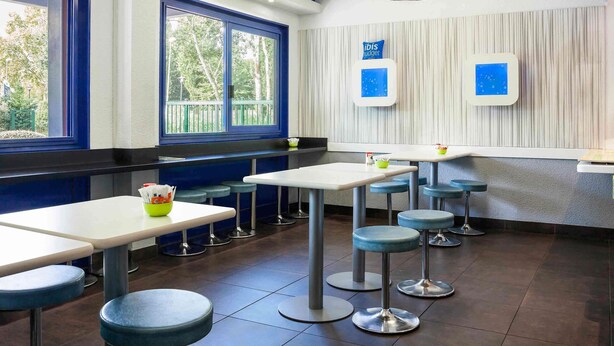 A modern restaurant interior with high tables and stools, large windows with blue frames, and a ceiling light fixture. - ibis budget Sucy en Brie (Sucy-en-Brie)
