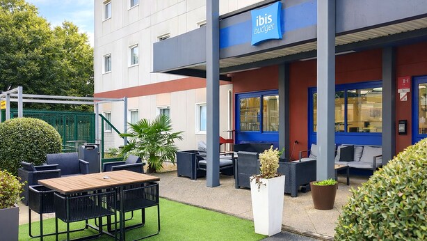 Un espace extérieur aménagé avec une table et des chaises, une plante en pot et un bâtiment sur lequel on peut lire l’inscription « ibis budget » sur une enseigne bleue.