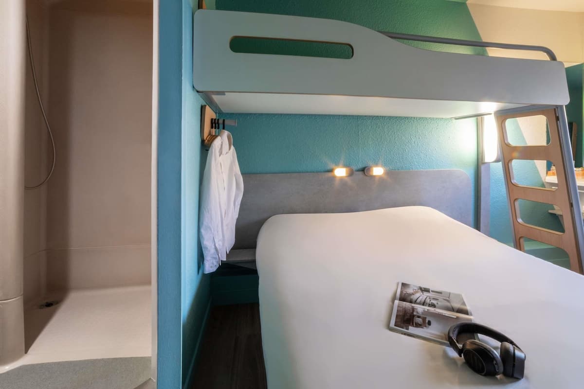 Un lit superposé avec un matelas blanc et une tête de lit dotée de deux lampes. Une robe blanche est accrochée sur le côté, et une paire d’écouteurs repose sur le lit.