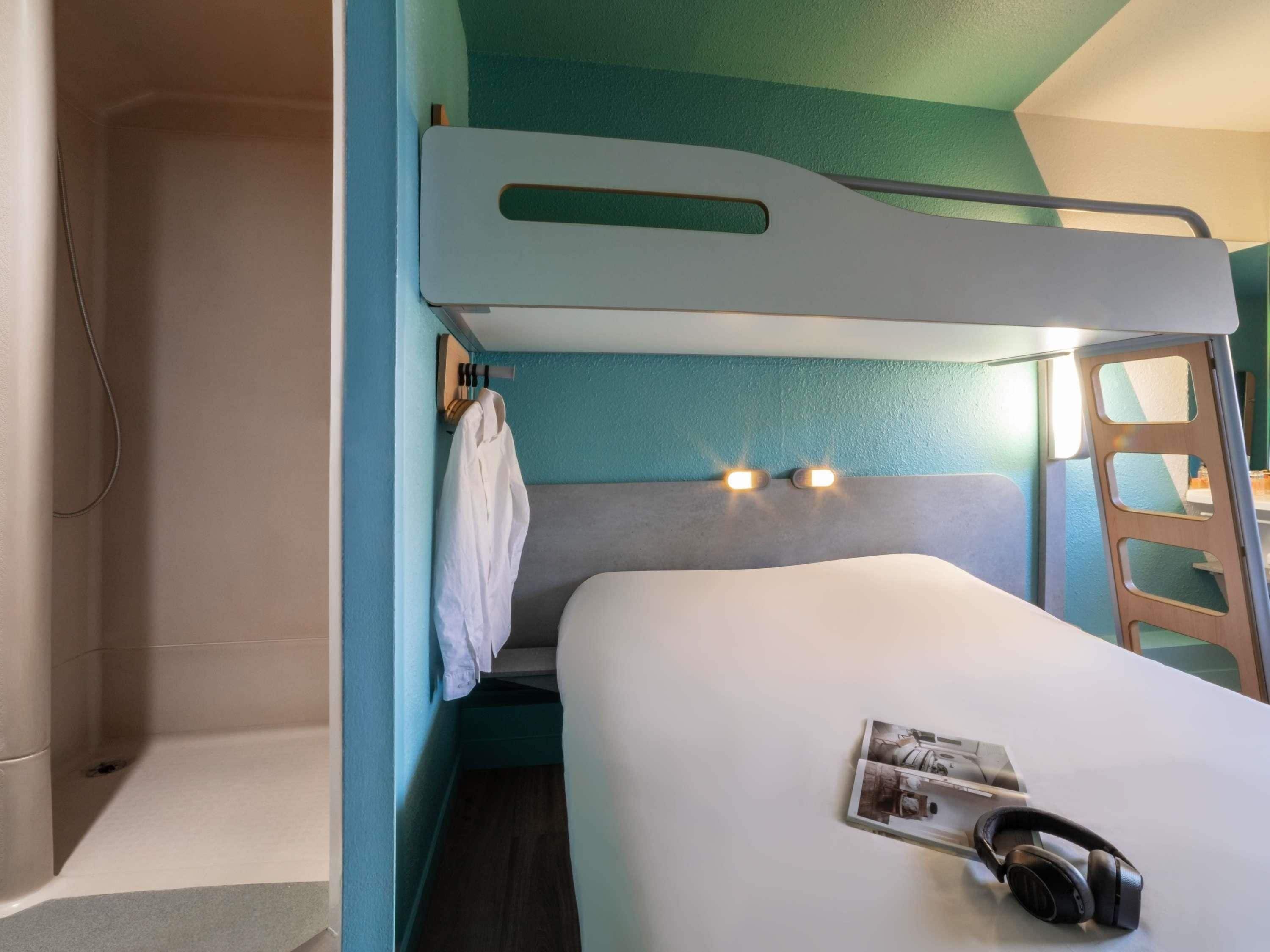 A bunk bed with a white mattress and a headboard with two light fixtures. A white robe hangs on the side, and a pair of headphones rests on the bed.