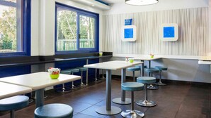 Een modern restaurantinterieur met hoge tafels en krukken, grote ramen met blauwe kozijnen en een plafondlamp.