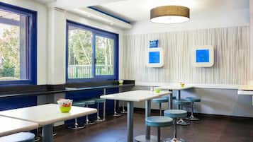 Een modern restaurantinterieur met hoge tafels en krukken, grote ramen met blauwe kozijnen en een plafondlamp.