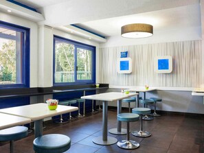 Ein modernes Restaurant mit hohen Tischen und Stühlen, großen Fenstern mit blauen Rahmen und einer Deckenleuchte.