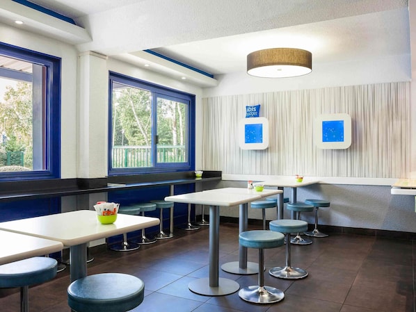 A modern restaurant interior with high tables and stools, large windows with blue frames, and a ceiling light fixture. - ibis budget Sucy en Brie (Sucy-en-Brie)