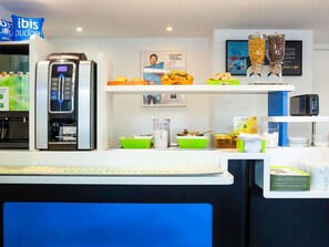 Un espace d’accueil d’hôtel avec une machine à café, des options pour le petit-déjeuner et un tableau encadré au mur.